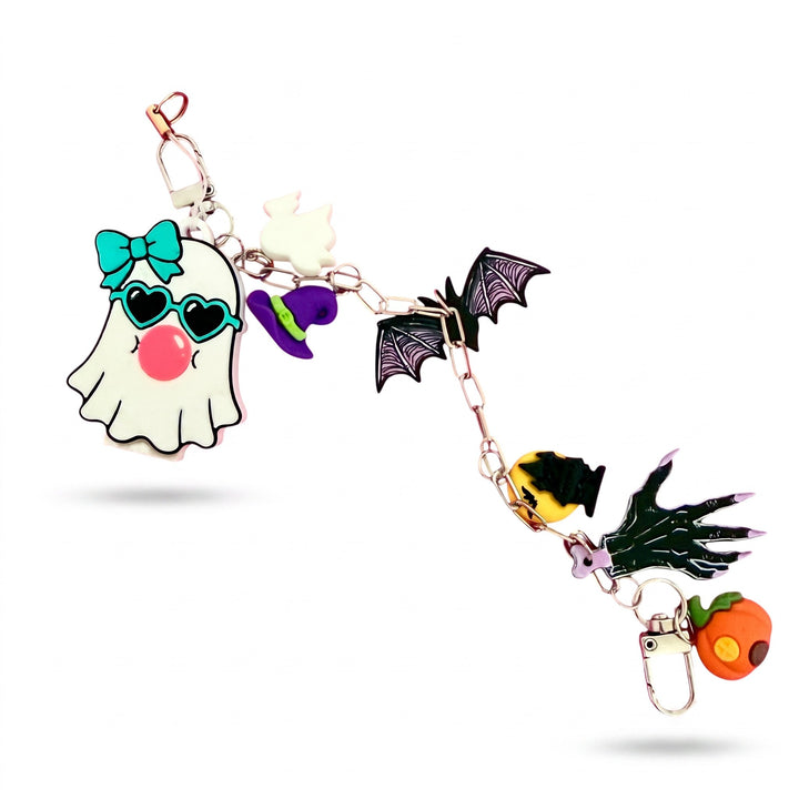 A Halloween-themed charm keychain featuring a white ghost with a teal bow and heart-shaped sunglasses, a purple witch hat, black bat wings, a yellow spider hand, and an orange pumpkin, all connected by rose gold-toned metal chains and clips.