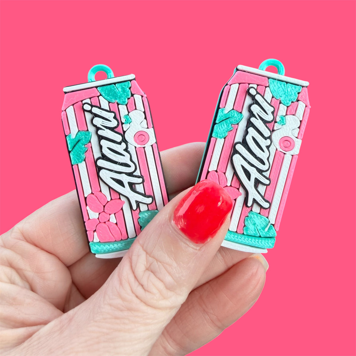 A hand with red nail polish holds two small aluminum can-shaped charms featuring the word 'Alani' in white script over pink and white vertical stripes, with teal and pink floral accents, against a solid pink background.