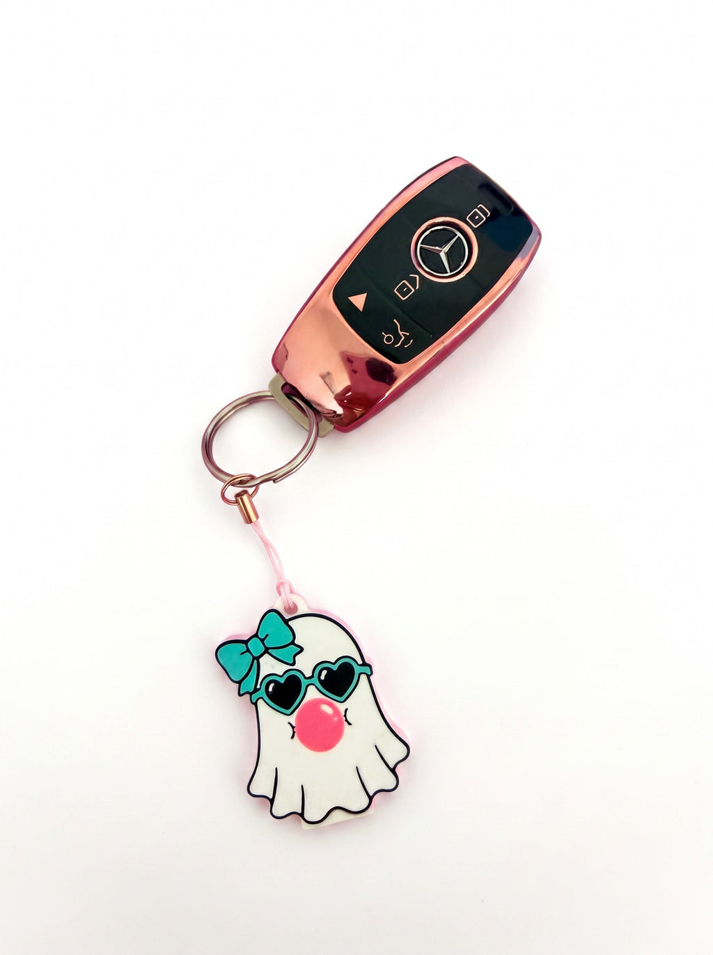A rose gold Mercedes-Benz key fob keychain with a pink metal key ring and a cute ghost charm wearing a teal bow, heart-shaped sunglasses, and a pink nose, shown on a plain white background.