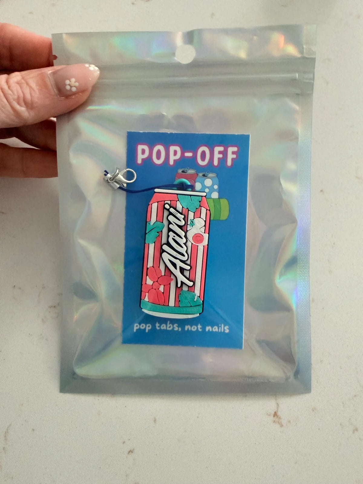 A hand holds a clear holographic zip pouch containing a pop-top accessory with a blue label that reads 'POP-OFF' and shows a colorful can illustration with the text 'pop tabs, not nails.'.