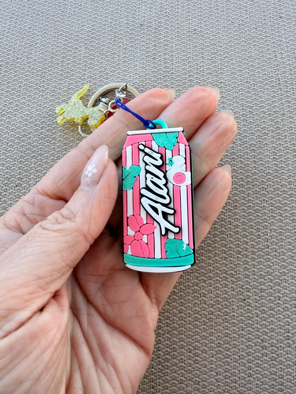 A hand holding a small Alani soda can keychain charm with pink and white stripes, floral accents, and a blue string attached, against a textured beige fabric background.
