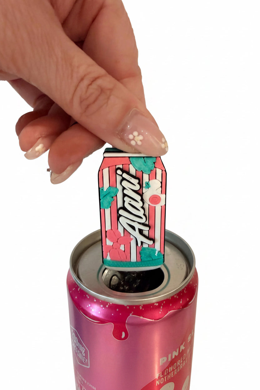 A hand with decorated nails holds a miniature Alani can-shaped keychain above an open pink beverage can, set against a white tiled wall background.