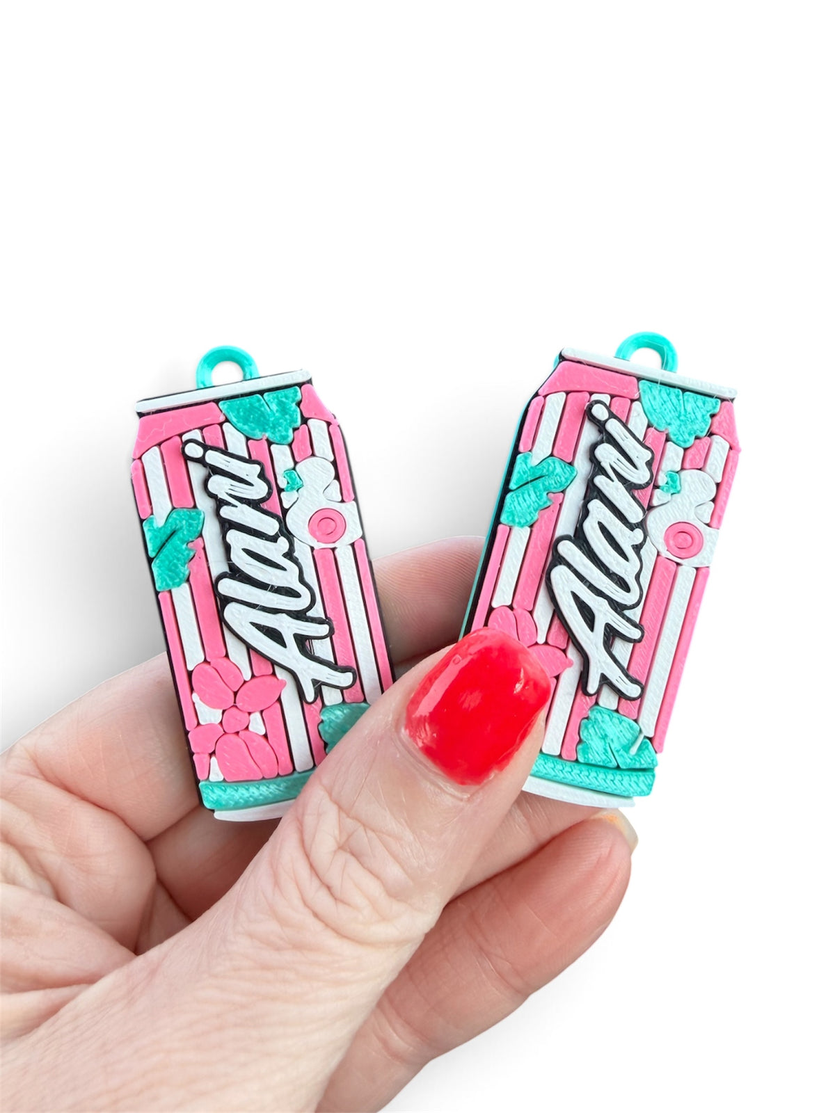 A hand with red nail polish holds two small pink and white striped can-shaped charms featuring the word 'Alani' in script and teal floral accents, against a plain white background.