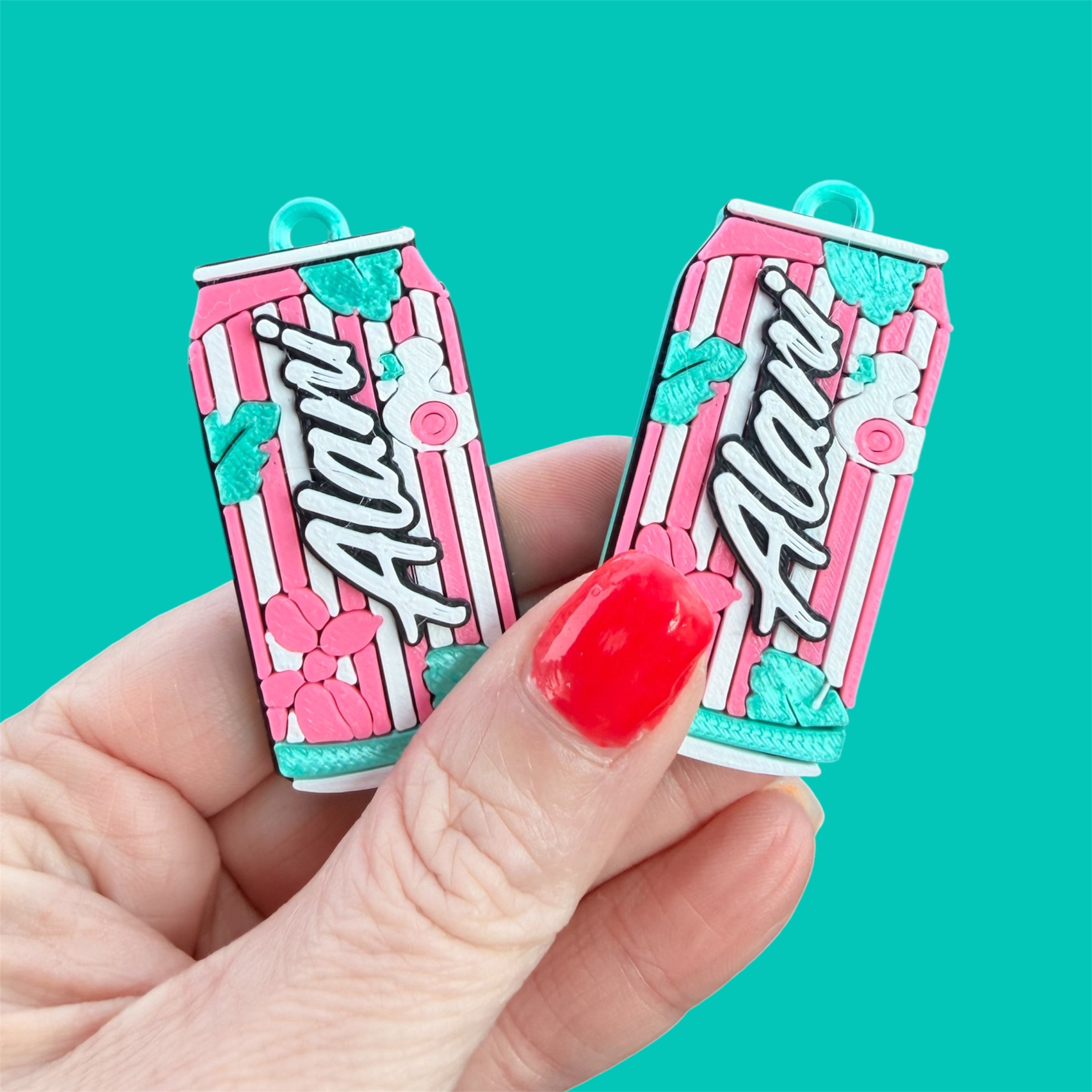 A hand with red painted fingernails holds two small aluminum can-shaped charms featuring pink and white vertical stripes, teal floral accents, and the word 'Alani' in white script on a bright teal background.