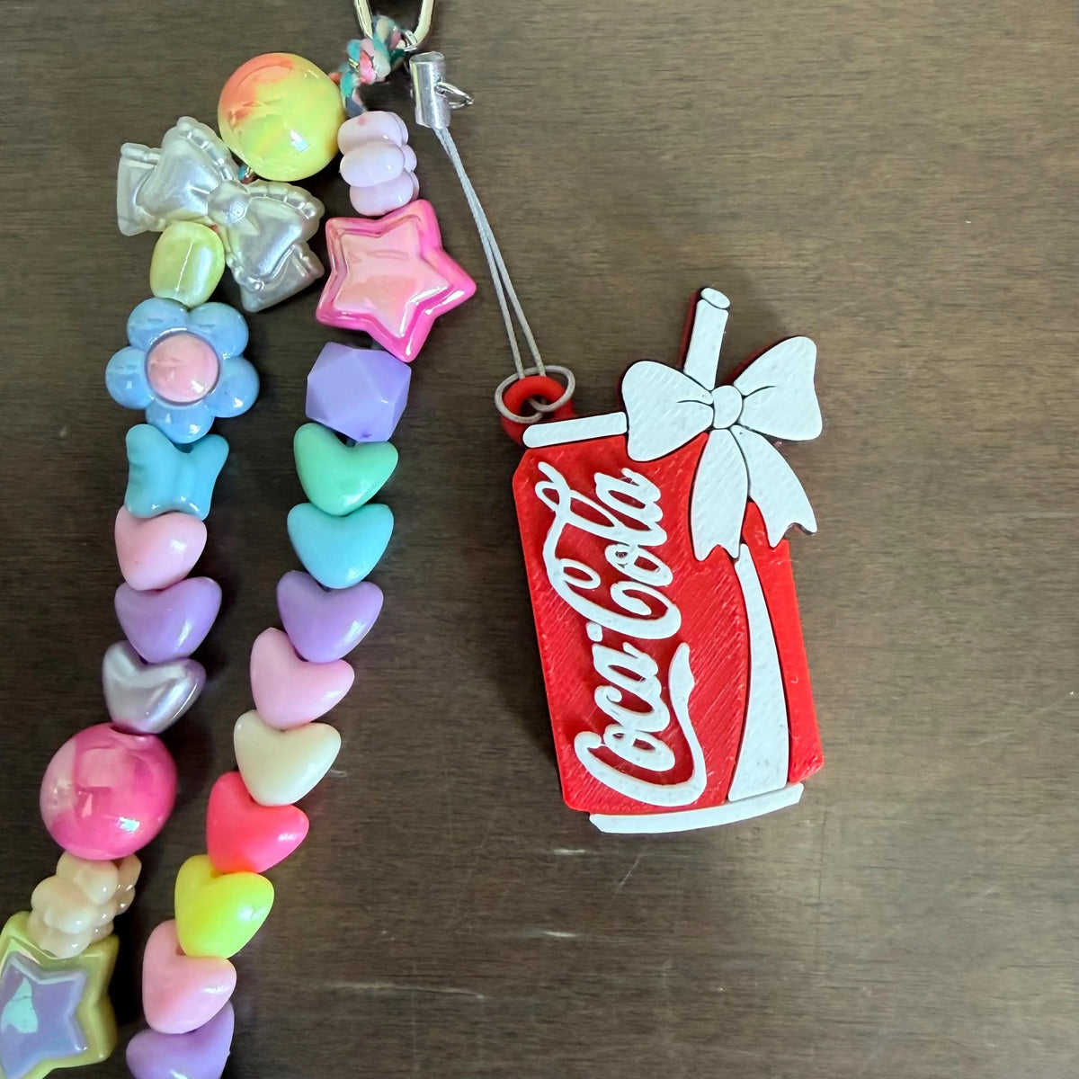 A handmade beaded charm keychain or phone strap featuring pastel heart and flower beads alongside a red Coca-Cola can-shaped charm with a white bow, displayed on a dark wooden surface.
