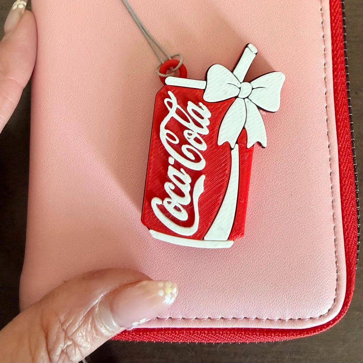 A close-up of a pink zip-around wallet with a red trim, featuring a red and white Coca-Cola can charm with a white bow attached near the top. A hand is holding the corner of the wallet.