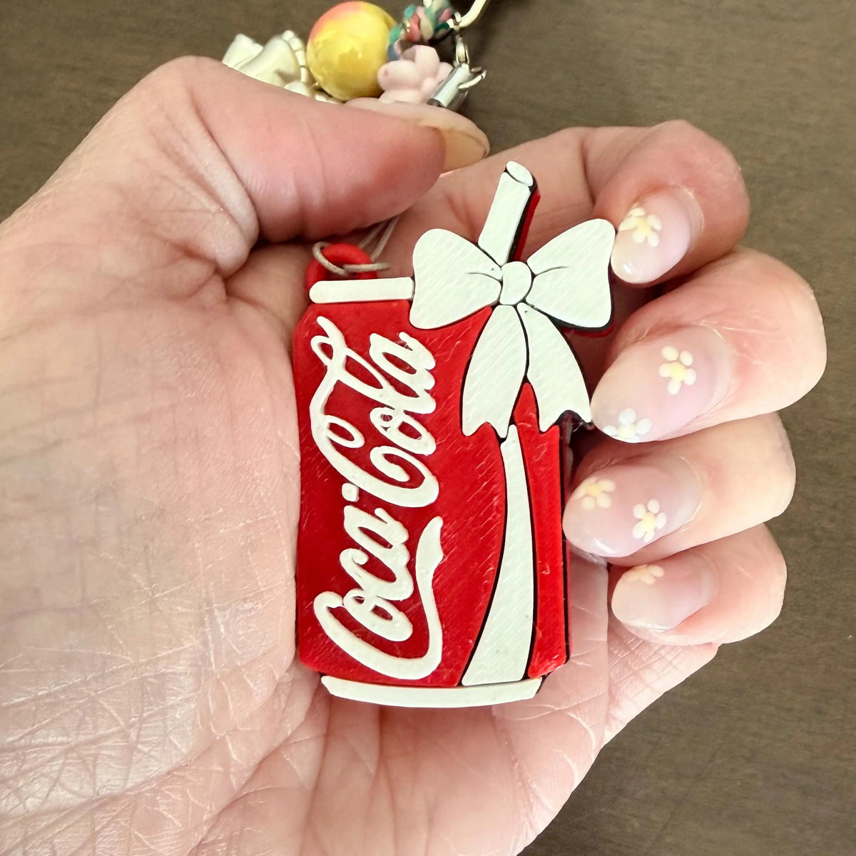 A hand holding a miniature red Coca-Cola can keychain charm with a white bow, shown against a neutral brown background.