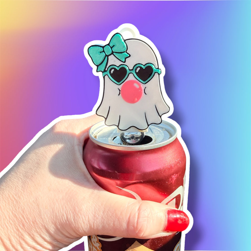 A hand with red nail polish holds a red aluminum can while inserting a ghost-shaped novelty bottle stopper. The stopper is white with a teal bow, black heart-shaped sunglasses, and a pink nose, shown against a colorful purple-to-blue gradient background.