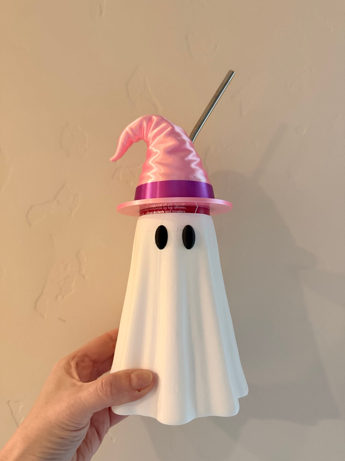 Reusable straw passing through the opening in a pink and purple witch hat topper on a ghost can koozie