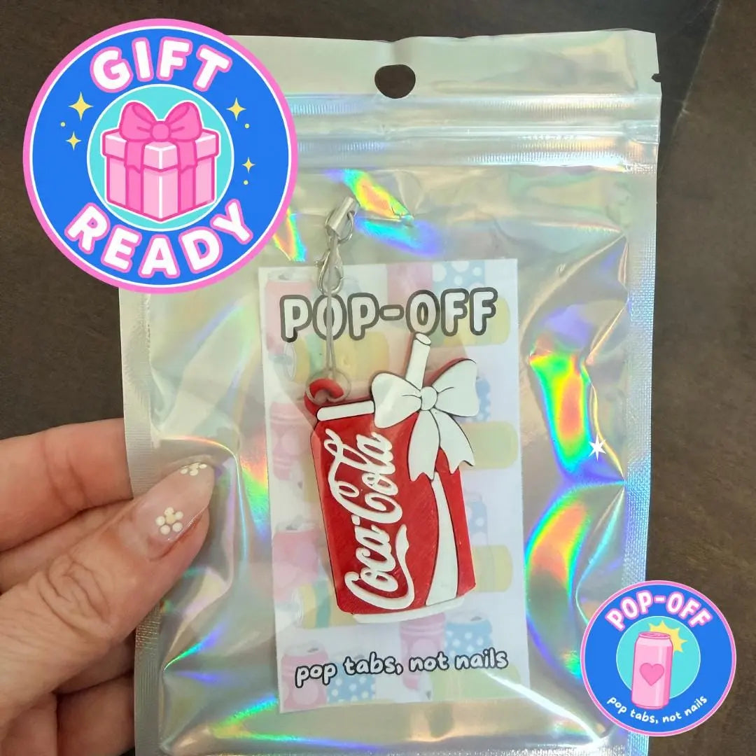 A hand holds a packaged novelty keychain shaped like a red and white Coca-Cola can with a white bow and straw. The item is sealed in a shiny holographic pouch with a printed insert reading 'POP-OFF' and 'pop tabs, not nails.