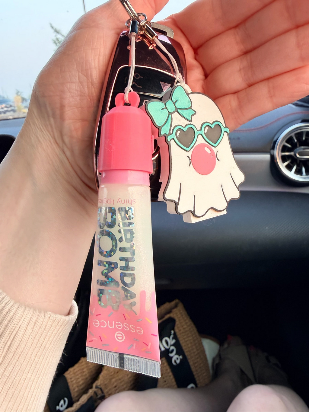 Pocket Magic essence Shiny Lipgloss and ghost keychain with heart sunglasses and bow, held in a hand inside a car.