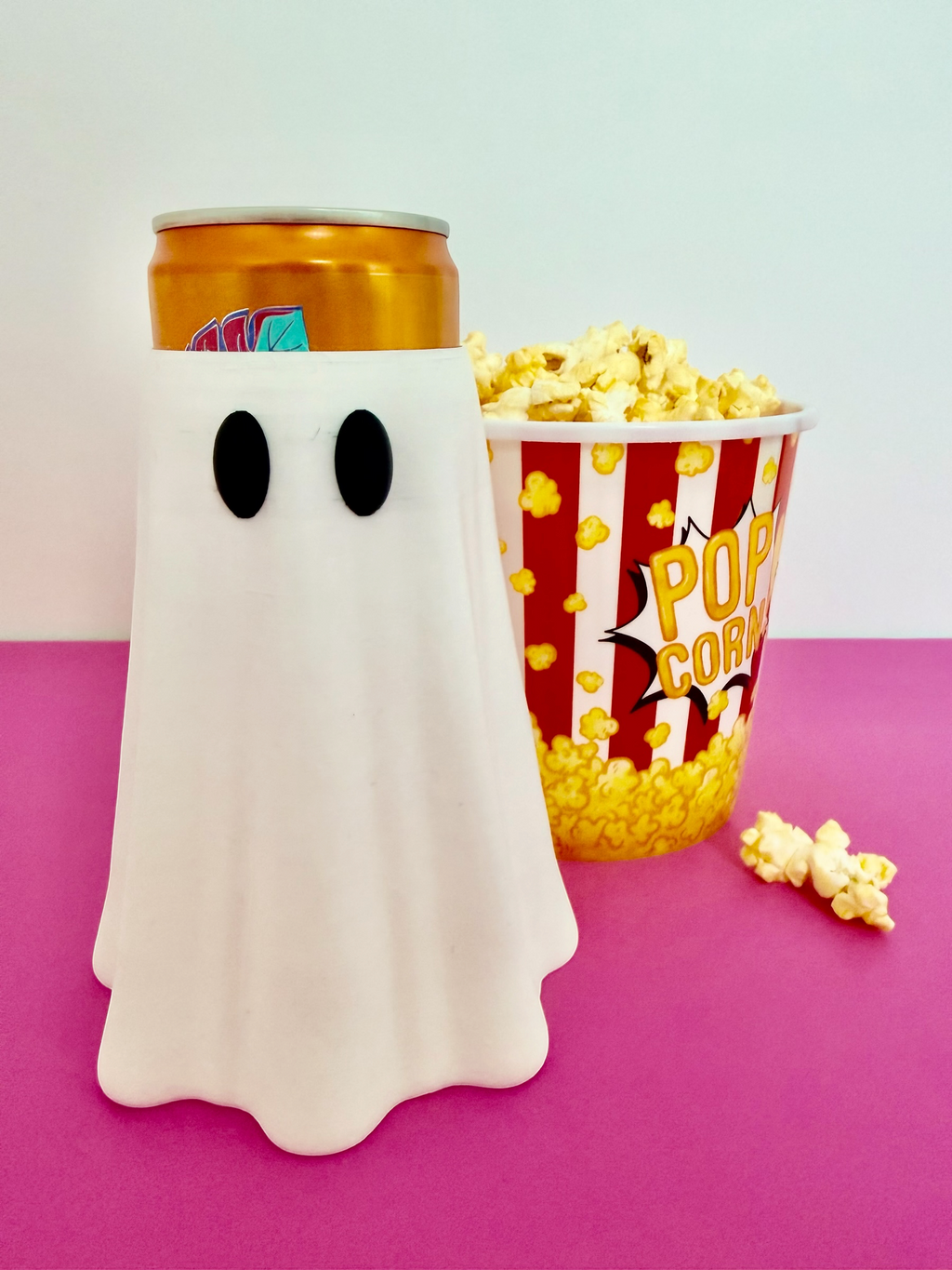 Pocket Magic white ghost insulated beverage can cooler sleeve next to a popcorn bucket on a pink surface
