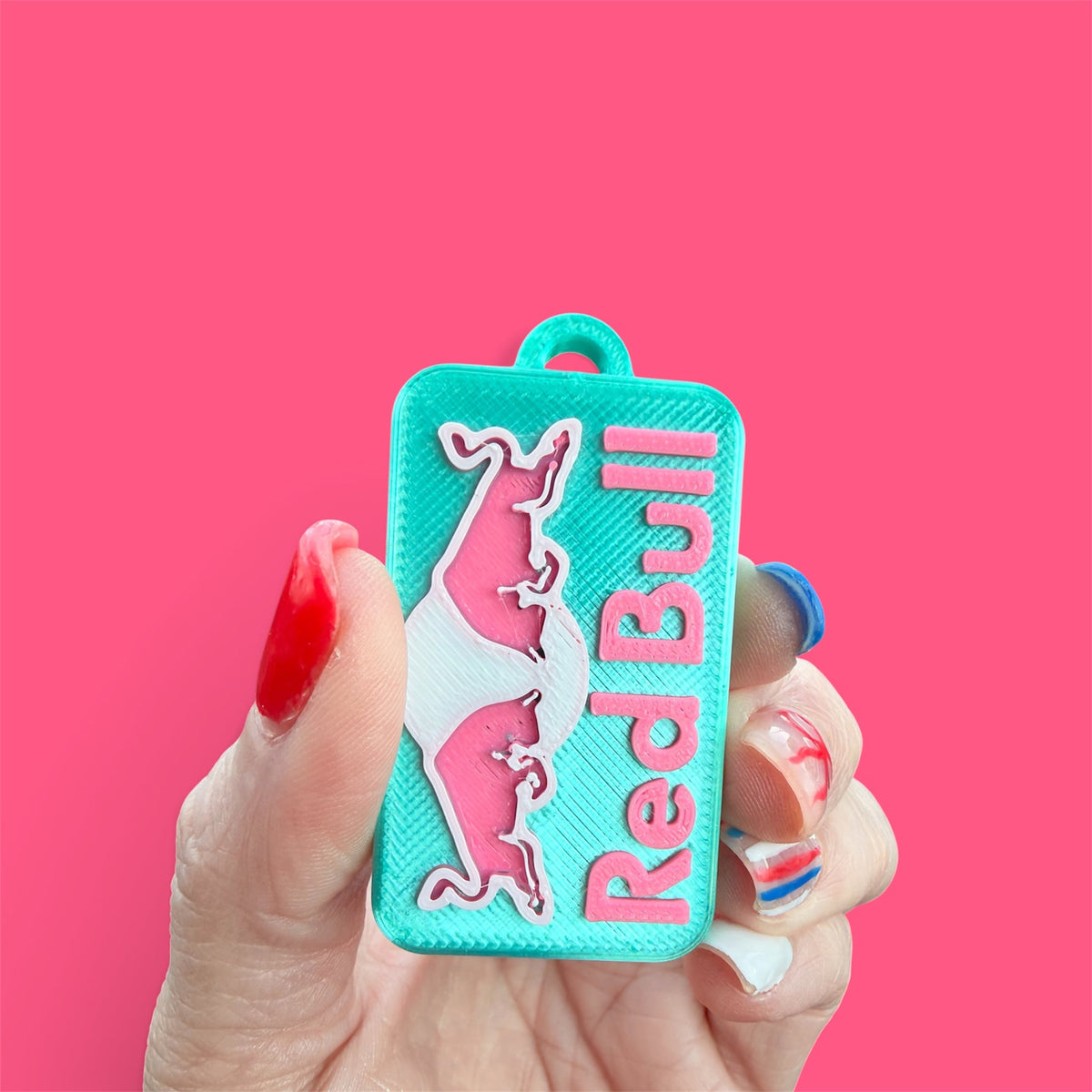 Teal Red Bull-inspired PopOff can tab opener keychain held against a pink background